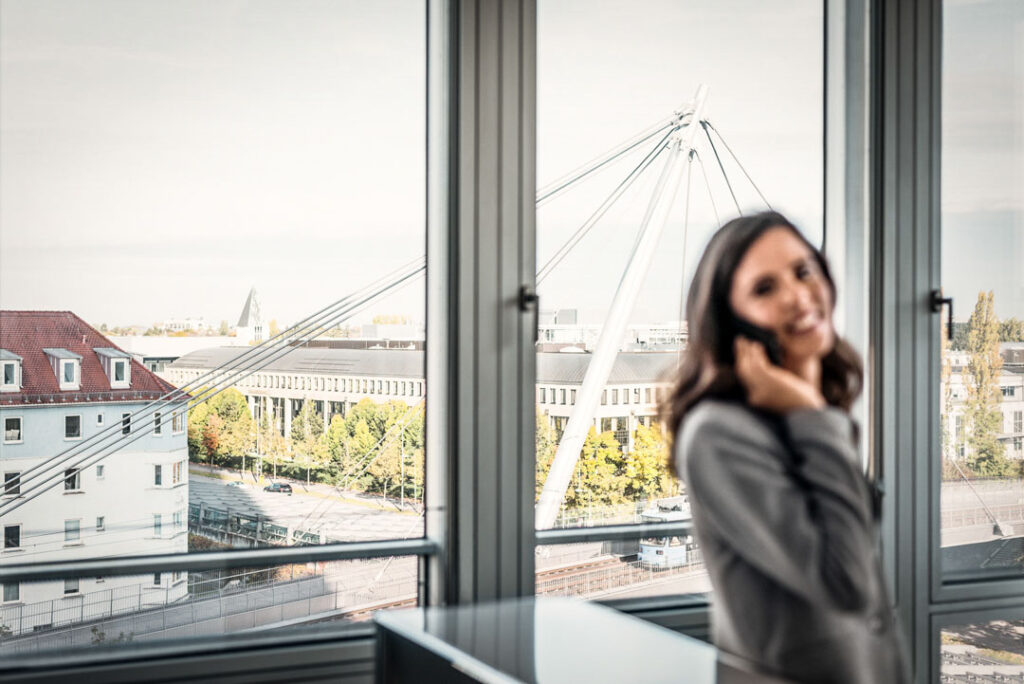 Geschäftsfrau telefoniert im Büro der MEAG AG in München, unscharfer Vordergrund, im Fokus die Fußgängerbrücke vor dem Fenster – Businessfotografie mit Blick auf Lebensqualität im Office.
