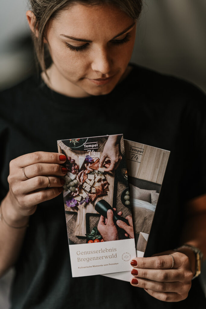 Junge Frau hält und präsentiert einen Hotelflyer in den Händen, freundlich lächelnd