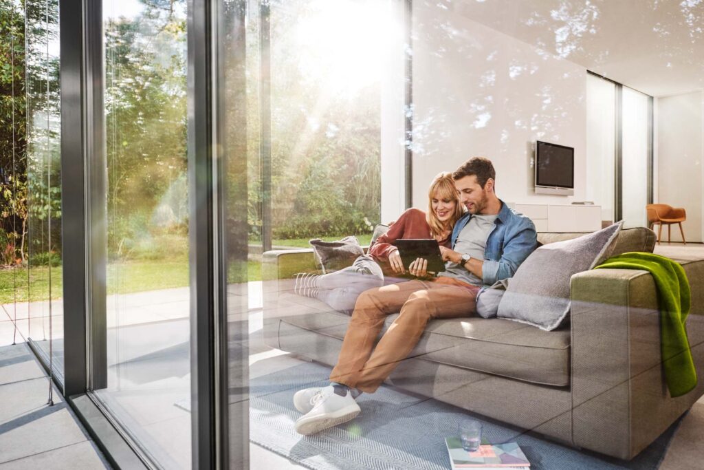 Paar sitzt entspannt auf einer Couch in einem designorientierten Architektenhaus und prüft gemeinsam Inhalte auf einem iPad – moderne Businessfotografie Hamburg für CosmosDirekt Versicherungen.