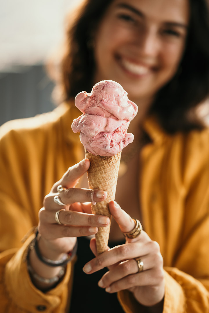 Dunkelhaarige Frau lacht unscharf in die Kamera und hält ein Erdbeereis in der Waffel, Fotografiert für Eismanufaktur von Businessfotograf