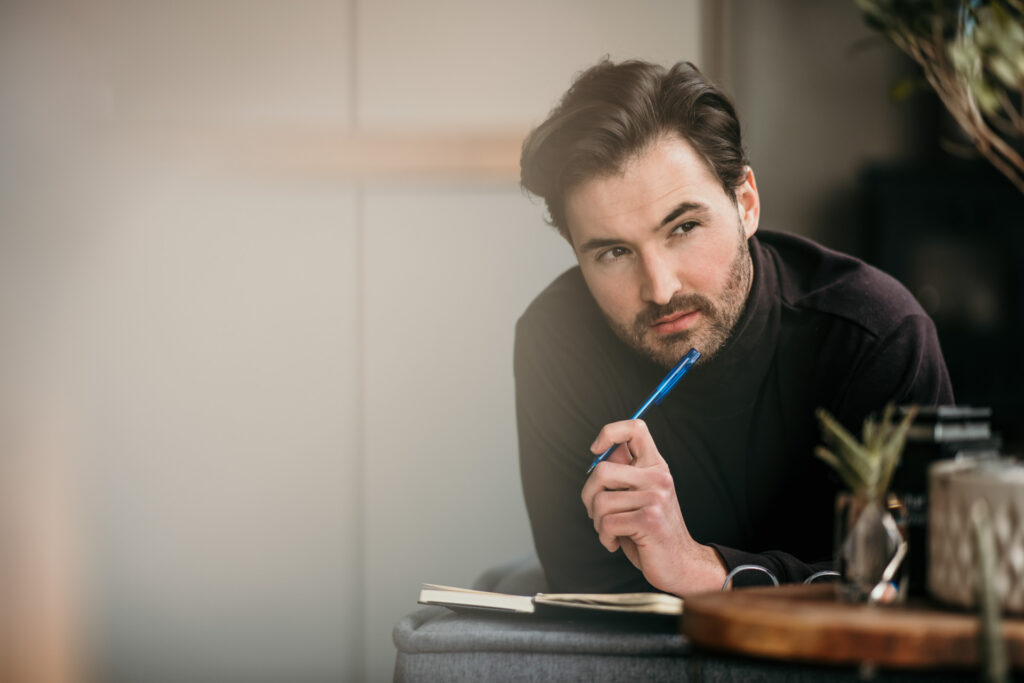 Junger Geschäftsmann liegt nachdenklich auf einer Couch, hält einen Stift in der Hand – authentisches businessfotografie hamburg-Portrait im modernen Büro.