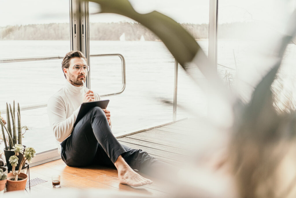 Junger Geschäftsmann sitzt nachdenklich am großen Fenster eines Hausbootes in Berlin, hält einen Stift in der Hand – businessfotografie.