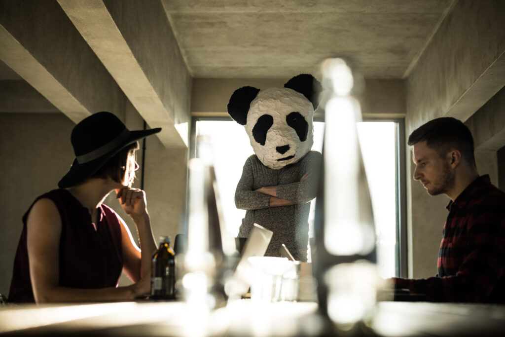 Zwei coole Office Workers sitzen an ihren Arbeitsplätzen, während eine Frau mit Panda-Maske zwischen ihnen steht und in die Kamera blickt – businessfotograf.