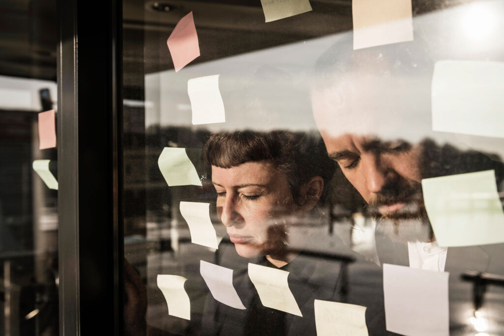 Erschöpfte Businesspeople lehnen ihre Stirn an eine Fensterscheibe, umgeben von bunten Post-its aus einem vorherigen Meeting – stimmungsvolle businessfotograf-Aufnahme im modernen Büro.