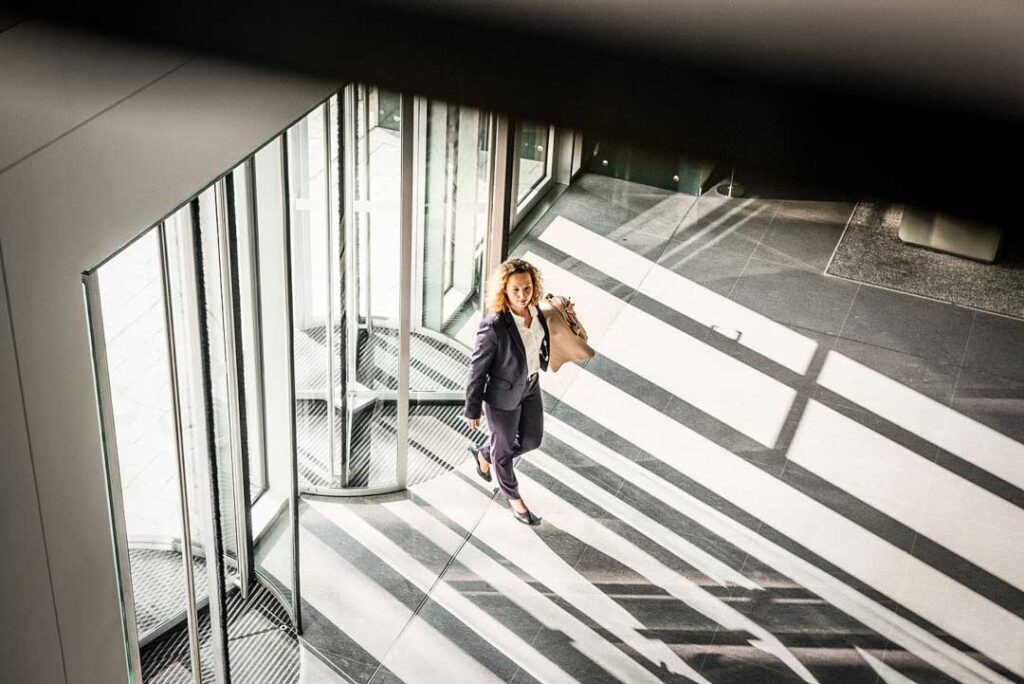 Businessfotografie zeigt eine Managerin im Anzug und Mantel, die energisch durch das Eingangstor der MEAG geht. Die Szene vermittelt Dynamik, Professionalität und den modernen Alltag von Führungskräften in einem Unternehmensumfeld.