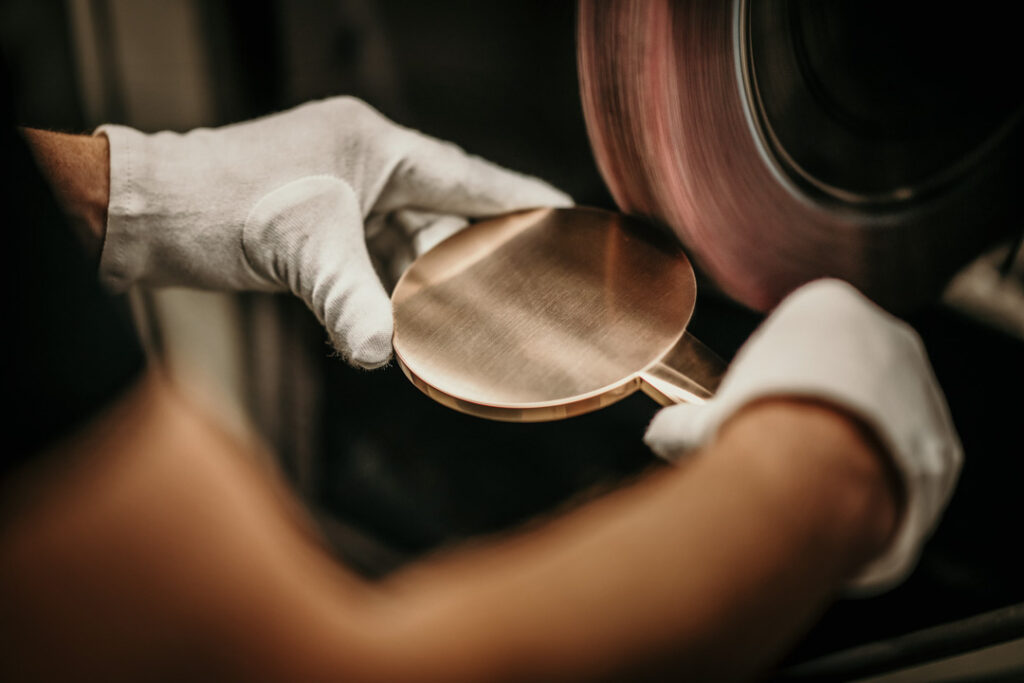 Hansgrohe Duschkopf in Bronze wird in der Fabrik an Poliermaschine von Händen mit weißen Handschuhen poliert – Close-up Business-Reportagebild, hochwertige Produktfotografie, aufgenommen von professionellem Businessfotografen und Werbefotografen.