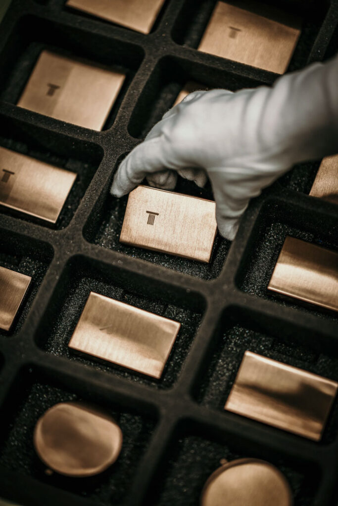 Dieses Bild zeigt die Fertigung in der Hansgrohe-Fabrik mit zahlreichen goldenen Knöpfen. Eine Hand mit weißem Handschuh nimmt einen Knopf heraus, wodurch Präzision, Qualität und Handwerkskunst symbolisiert werden. Das Motiv eignet sich ideal für Business-Fotos, Unternehmenskommunikation, Kampagnen und Social Media, die höchste Produktionsstandards, Premium-Qualität und Markenwert vermitteln. Die stilvolle, detailreiche Bildsprache entspricht dem Stil eines professionellen Werbefotografen aus Berlin und unterstreicht Exzellenz, Handwerkskunst und Vertrauen in die Marke Hansgrohe. Fotografiert von professionellem Businessfotografen