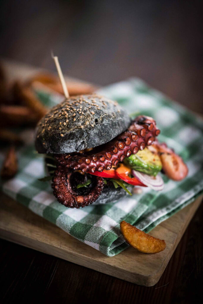 Food Fotografie zeigt einen Oktopus-Arm in einem Burger Bun, angerichtet als Oktopus-Hamburger und servierfertig – Foodfotograf hält die kreative Präsentation fest, Food Fotograf inszeniert das Gericht, Foodfotograf Hamburg und Foodfotograf Berlin dokumentieren die außergewöhnliche Speise.