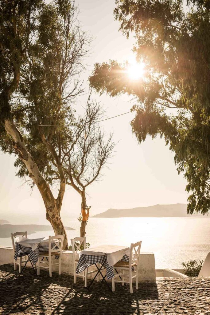 Restaurant Marketing Agency zeigt Tische mit herrlicher Aussicht in der Sonne auf Santorini, Griechenland – stilvolle Präsentation für Restaurantmarketing und Food-Erlebnis in Fira.
