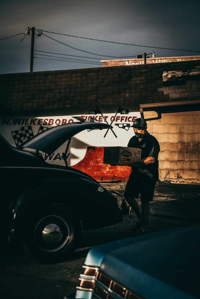 Foodfotograf zeigt Brian Call, wie er Moonshine in den Kofferraum eines alten Schmugglerfahrzeugs lädt – Call Family Distillery, Wilkesboro, North Carolina, USA, mit historischem Bezug zur NASCAR und der Moonshine-Familiengeschichte.