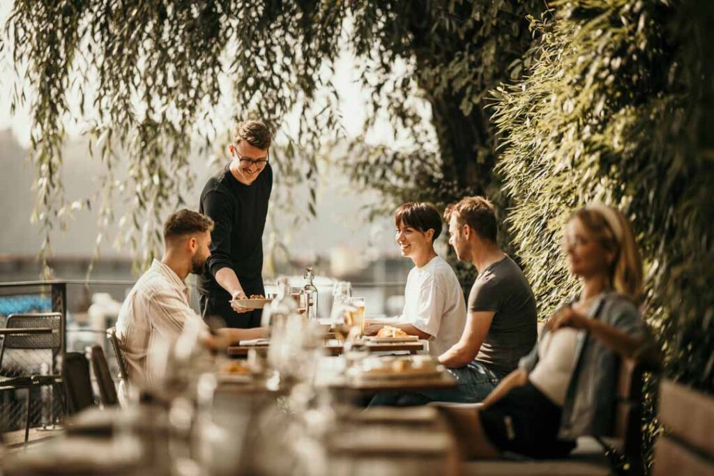 Gäste werden auf der Terrasse der Hafenküche Berlin bedient, lachender Kellner sorgt für lebendige Atmosphäre – professionelle Hospitality-Fotografie für Restaurantkommunikation.