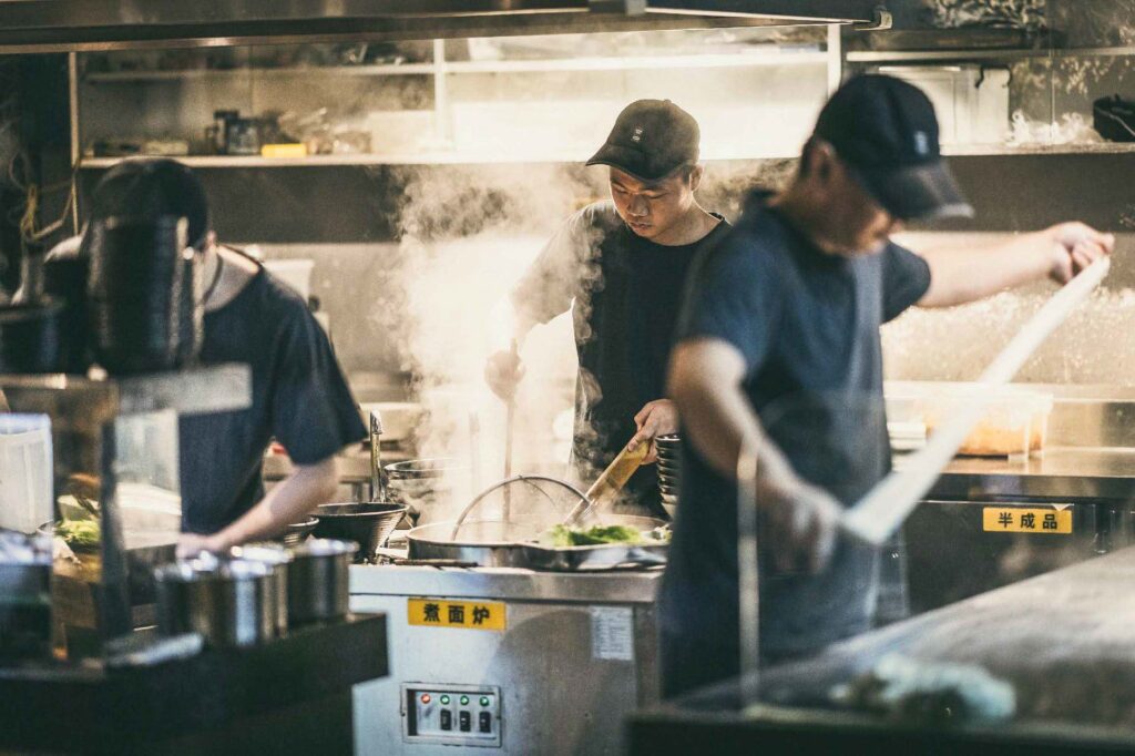 Drei Köche in einer chinesischen Restaurantküche ziehen handgemachte Nudeln auseinander, während Dampf aus den Töpfen steigt – dynamisches Action-Motiv für Food- und Restaurantfotografie.