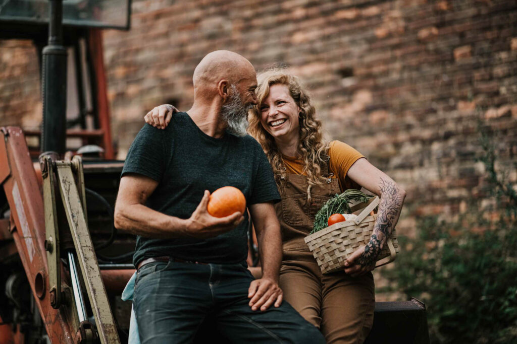Foodfotograf: Junge Biobäuerin und älterer Kollege lachen gemeinsam am Traktor, während sie frisch geerntetes Gemüse auf dem Biohof genießen.