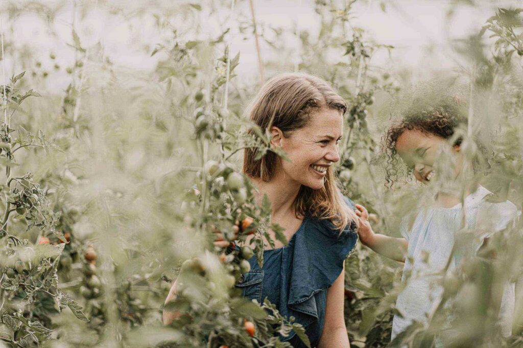 Foodfotografie zeigt Mutter und Tochter, die sich lachend in einem sonnigen Tomatenfeld ansehen.