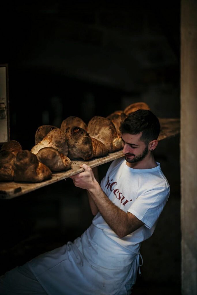 Food Fotograf München zeigt einen italienischen Bäcker, der traditionelles Brot aus dem Holzfeuerbackofen auf einem speziellen Tablett holt – handwerkliche Backkunst im Detail.