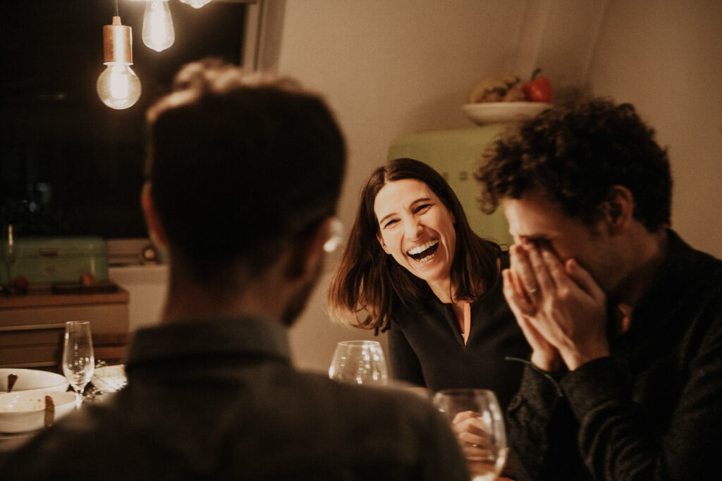 Foodfotograf zeigt eine Frau, die ihre Freunde am Esstisch lachend anschaut – stimmungsvolle Abendatmosphäre.