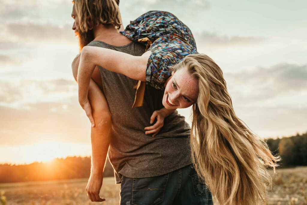 Ein Mann trägt eine Frau auf dem Rücken, während sie in der Natur unterwegs sind. Frau Lacht herzlich.