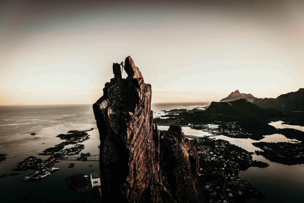 Influencer Chris Burkard steht zwischen zwei Gipfeln auf den Lofoten, mit zerklüfteten Buchten unterhalb und Sonnenuntergang am Horizont – episches Bergsteiger- und Reisemotiv für die Lufthansa-Kampagne ‚Life Changing Places‘ von Accenture Song. Fotografiert von Werbefotograf Berlin.