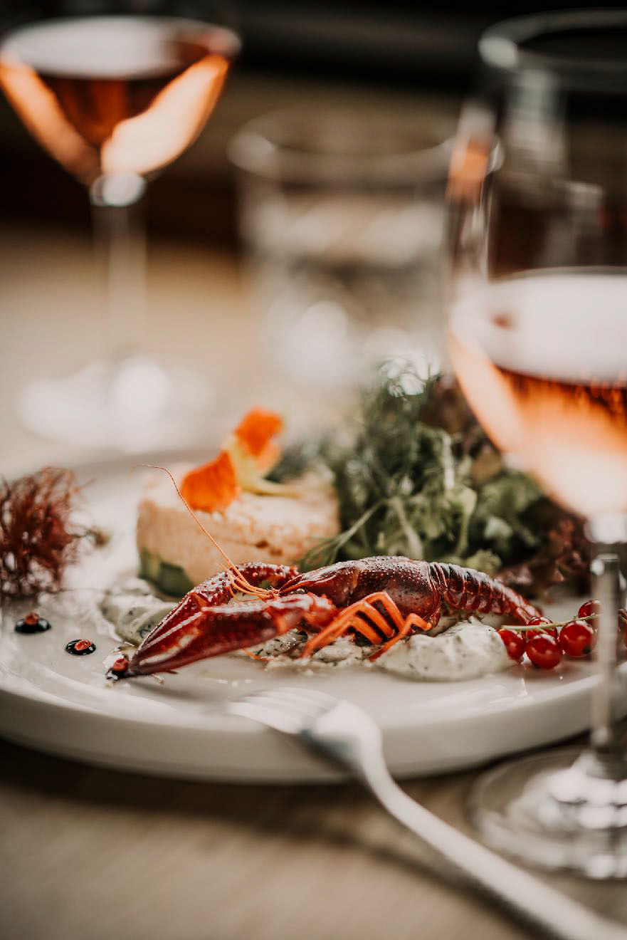Flusskrebs-Hummer-Foodfotograf-berlin Tellergericht mit rotem Flusskrebs oder Hummer, umgeben von rosafarbenen Weingläsern, viel Unschärfe – stilvolles Food-Foto mit Premium-Atmosphäre.