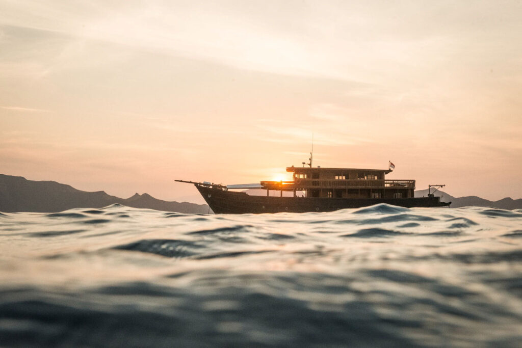 Ein Schiff fährt auf ruhigem Wasser während eines Sonnenuntergangs mit sanften Wellen im Vordergrund. Kreuzfahrtschiff Amanikan, Aman Hotels