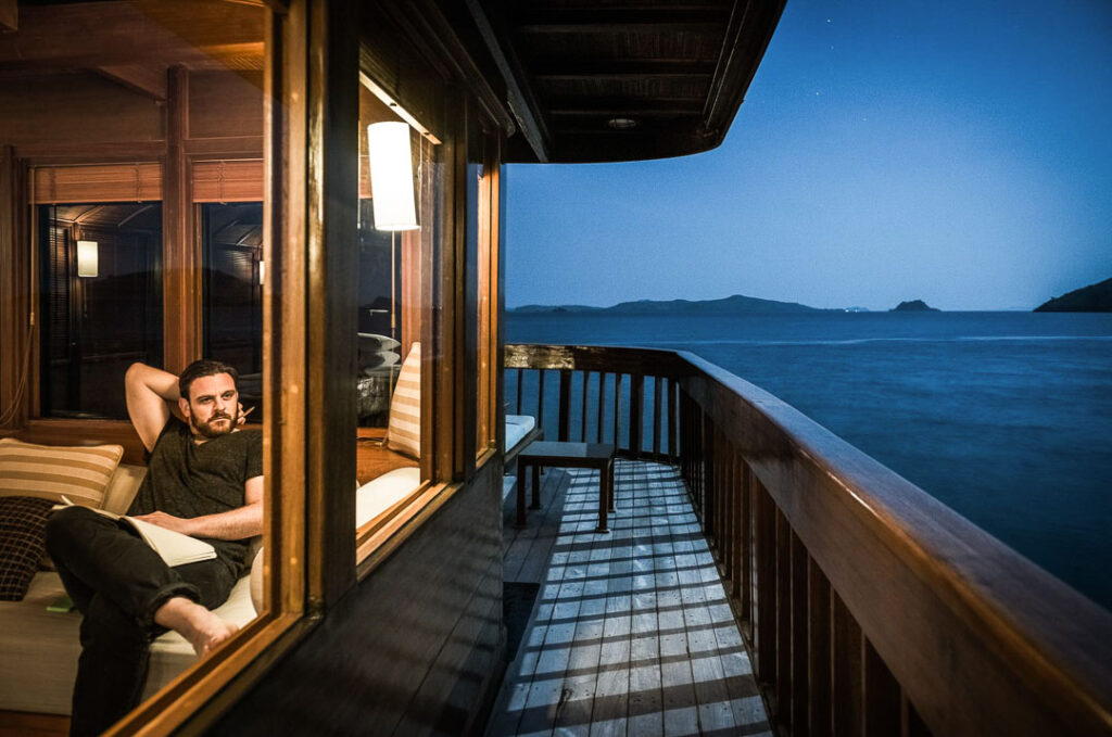 Gast auf dem Privat-Kreuzfahrtschiff Amanikan schaut bei Nacht aus der Kabinenfenster, stimmungsvolle Hotelfotografie für Aman Hotels und Lufthansa aufgenommen von einem Hospitality Fotograf.