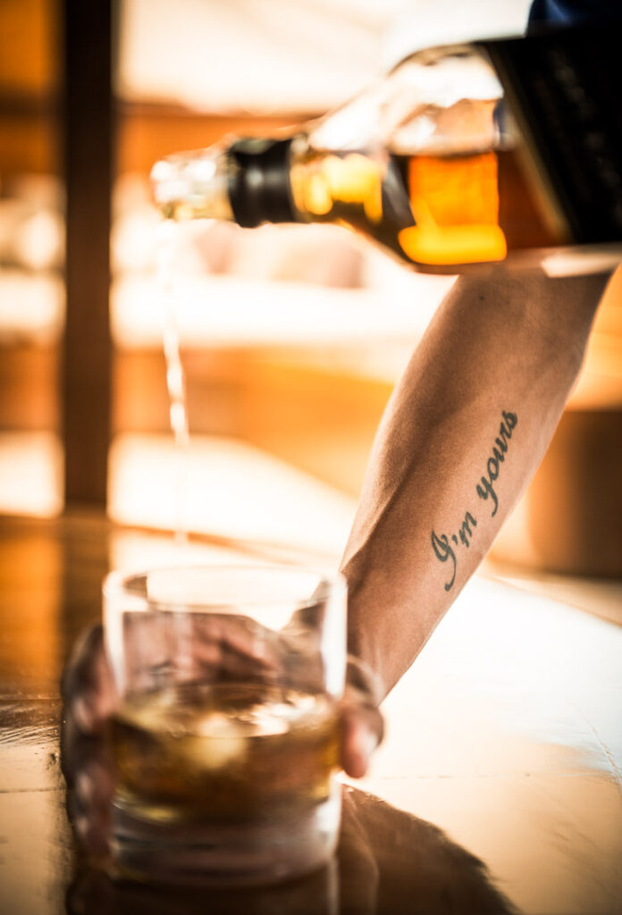 Arme gießen edlen Whiskey in Glas, Tattoo ‚I’m yours‘ auf Arm sichtbar – Hotelfotografie, stilvolle Baraufnahme.