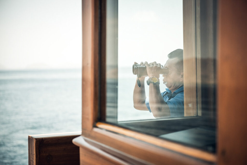 Kapitän der Amanikan schaut durch ein Fernglas aufs Meer, stimmungsvolle Hotelfotografie für Aman Hotels und Lufthansa festgehalten von einem Hotel Marketing Fotograf.