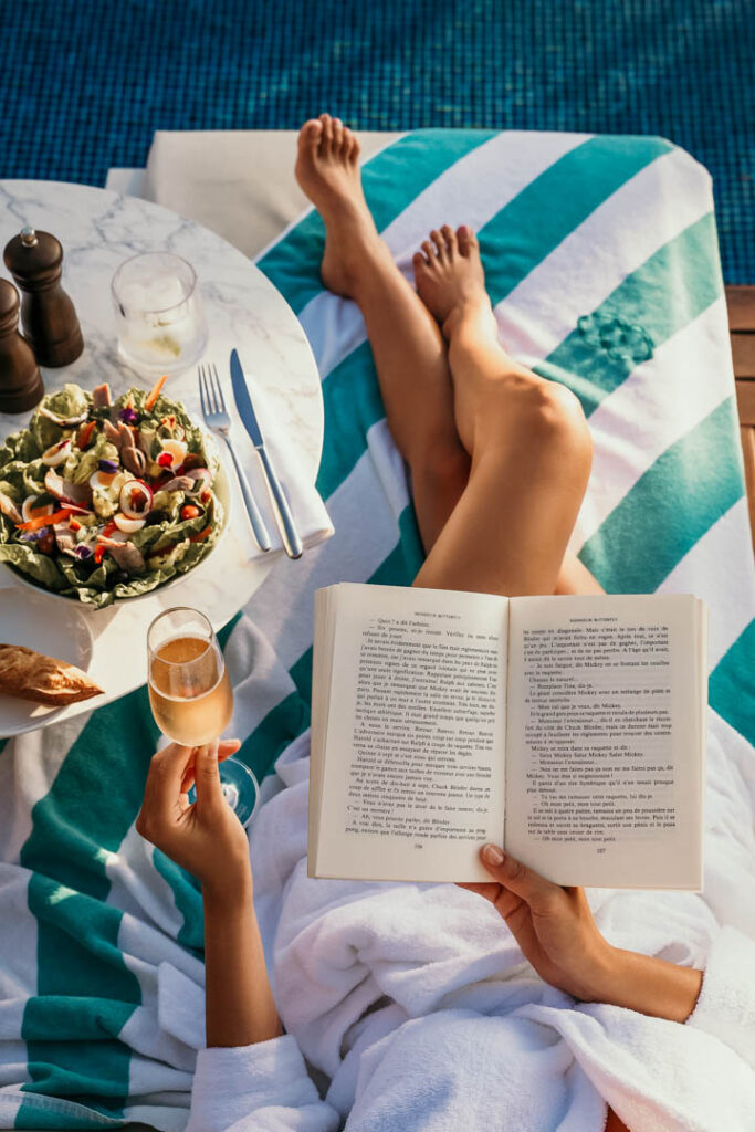 Frau im Bademantel entspannt auf Liegestuhl am Sofitel-Pool, hält Buch und Glas Champagner, daneben Salat Niçoise auf Tischchen – Werbefotograf für Hospitality-Marketing