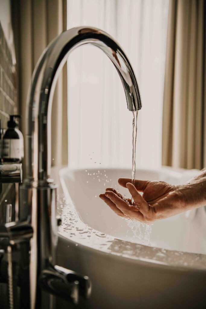 „Männliche Hand fängt Wasser aus Axxor Badewanneneinlauf über freistehender Badewanne, Close-up, Wasser spritzt – luxuriöses Werbefoto im Hotel VivaMayr Altausee.“