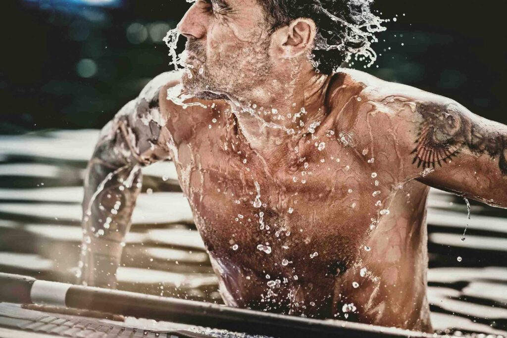 „Mann stützt sich auf seinem SUP im Wasser auf und schüttelt energisch das Wasser vom Kopf – sportliches Actionmotiv mit dynamischem Wassersplash.“