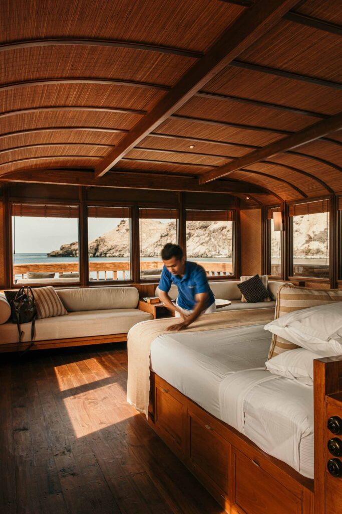 Innenraum des Privat-Charter-Kreuzfahrtschiffes Amanikan, klassisch-holzig eingerichtetes Hotelzimmer, während ein Mitarbeiter das Bett macht – hochwertiges Hospitality-Foto für die Hotelgruppe Aman. Hotelfotografie, Werbefotografie