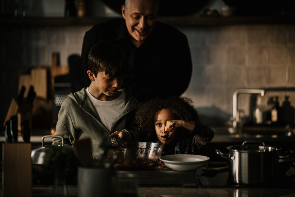 Alexander Herrmann lässt Kinder beim Kochen probieren, lebendige Szene in dunkler Küche, authentisch eingefangen von einem spezialisierten Foodfotografen.