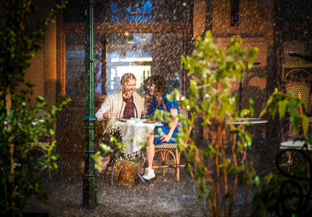Paar lacht an Esstisch in Kaffee während Regenguss, fotografiert von Werbefotograf auf Werbereh für einen TVC.