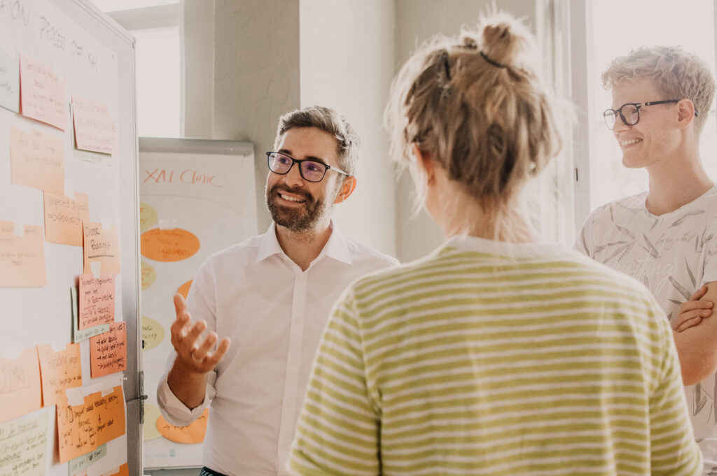 Drei Teilnehmer eines Businessmeetings stehen am Flipchart und diskutieren angeregt miteinander, authentische Szene eines Businessportraits