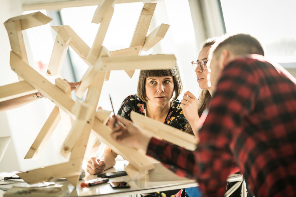 Drei MitarbeiterInnen halten ein kreatives Meeting an einer Holzskulptur in modernem Büro – businessfotografie.