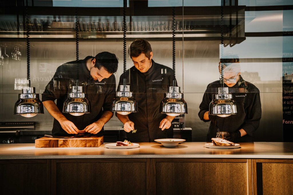 Drei Köche arbeiten konzentriert in der offenen Küche des Restaurants Hafenküche Berlin – lebendige Szenerie, festgehalten in moderner Hospitality-Fotografie für starke visuelle Restaurantkommunikation.