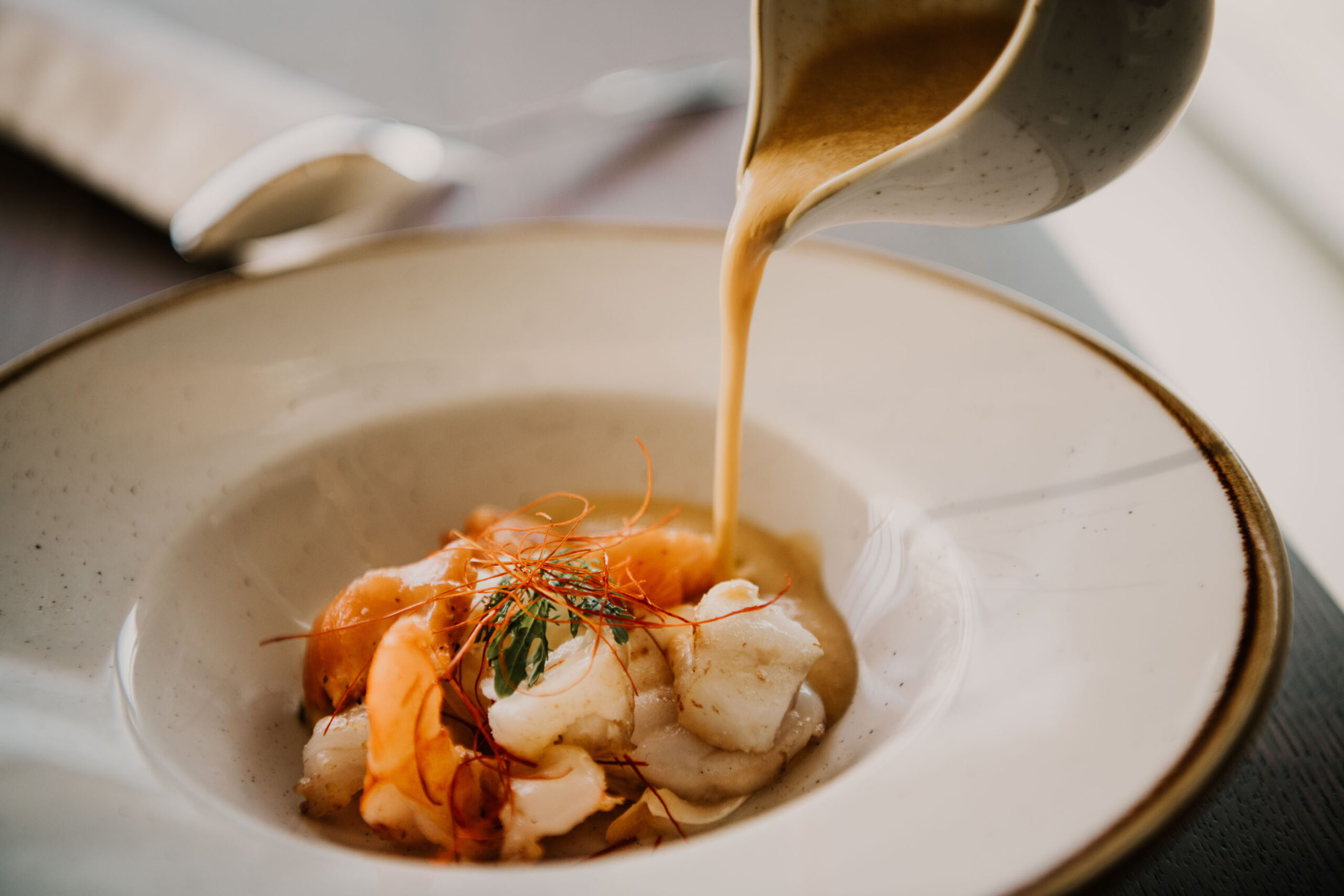 Fischgericht auf modernem Teller, Krabbensoße wird aus Soßiere übergegossen – hochwertiges Food-Foto für Hafenküche Berlin.