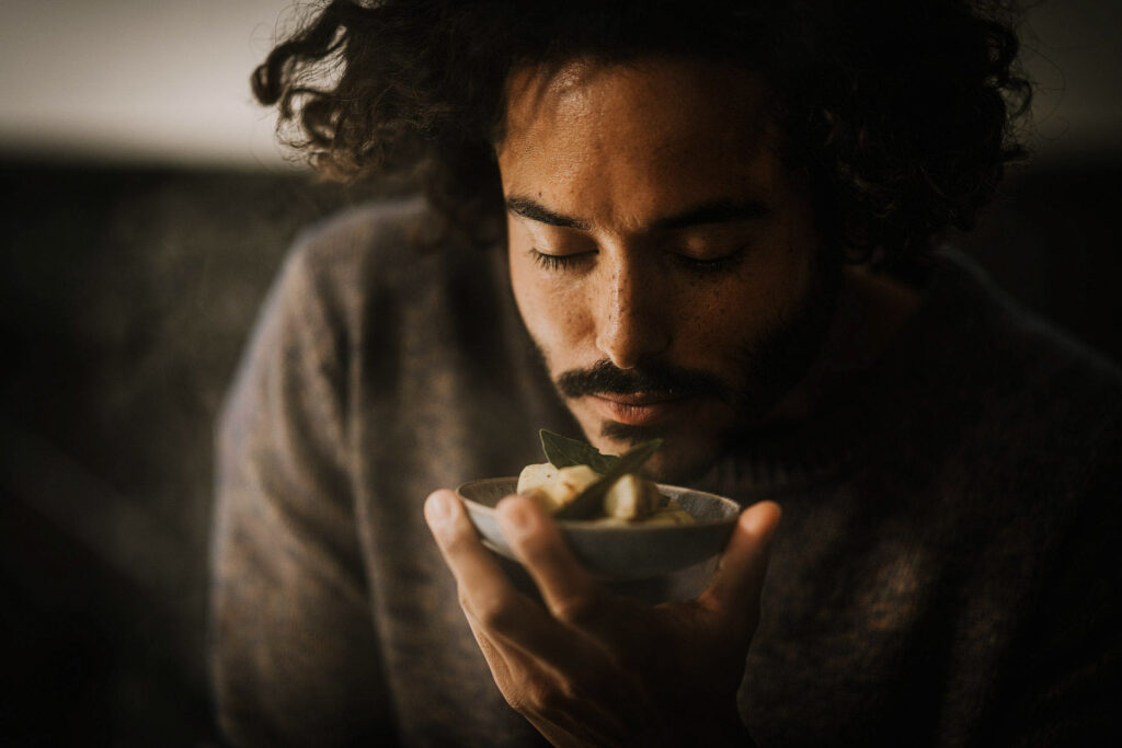 „Schöner Mixed-Race-Mann mit Sommersprossen und Afro-Locken riecht genießerisch mit geschlossenen Augen an einem warmen Vorspeisenteller – stimmungsvolles Werbemotiv für Fissler.“