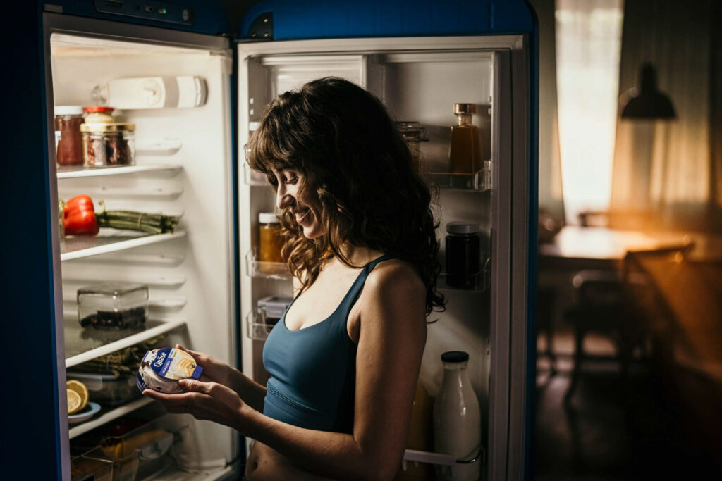 Food Fotografie zeigt eine junge Frau, die eine Packung Quäse aus ihrem Kühlschrank in der Küche holt – Foodfotograf fängt den Alltagmoment ein, Food Fotograf inszeniert die Szene, Foodfotograf Hamburg und Foodfotograf Berlin dokumentieren das authentische Setting.