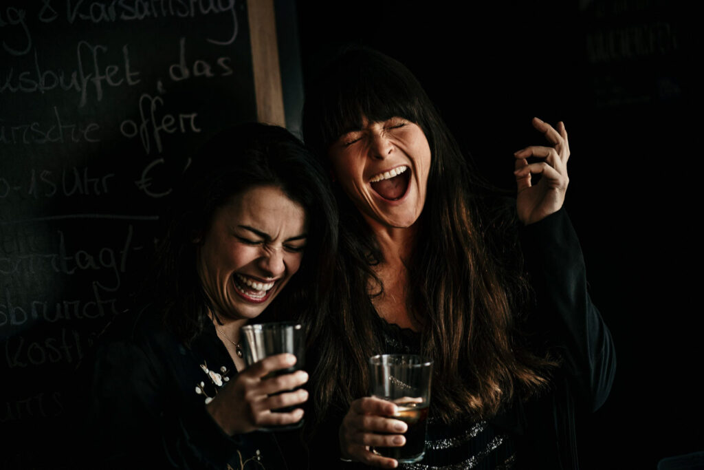 Food Fotografie zeigt Freundinnen, die herzlich lachen, nachdem sie ein Glas Cola auf Ex getrunken haben – die Kohlensäure kitzelt in der Nase.