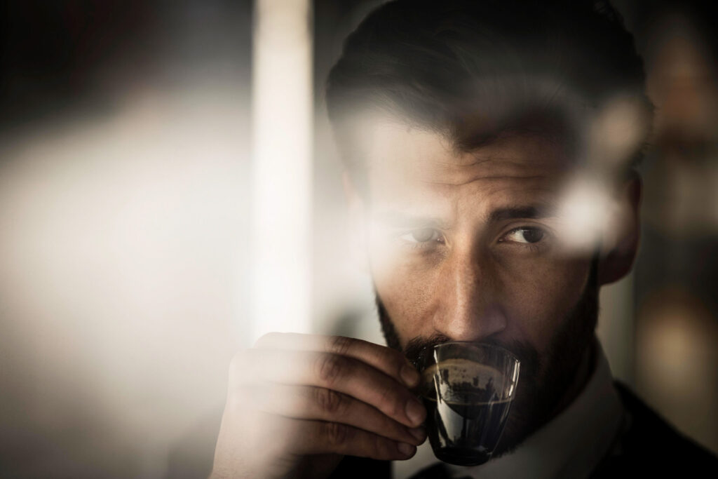 Hübscher Mann trinkt Espresso aus einer Tasse, fotografiert durch ein Fenster mit Spiegelungen – stimmungsvolles Portrait für Lifestyle- und Werbefotografie.
