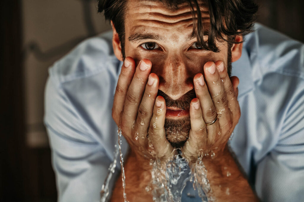 Mann wäscht sich das Gesicht, Wasser läuft über sein Gesicht, beide Hände nah am Gesicht, Blick in den Spiegel – Werbefotografie, Werbefotograf Berlin.