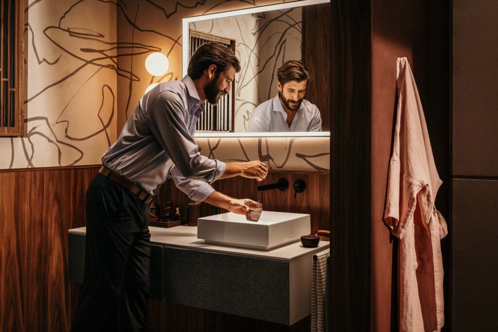 Mann zapft Wasser aus Hansgrohe Armatur in Matt-Schwarz in dunkelrotem asiatisch inspiriertem Badezimmer – Werbefotografie, Werbefotograf Berlin.