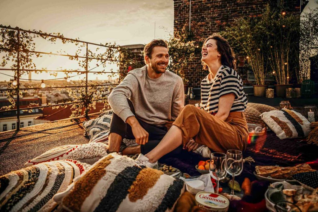 „Paar lacht auf Picknickdecke im Sonnenaufgang, mit Geramont-Käse – authentische Lifestyle- und Food-Fotos für Genussmomente im Freien.“