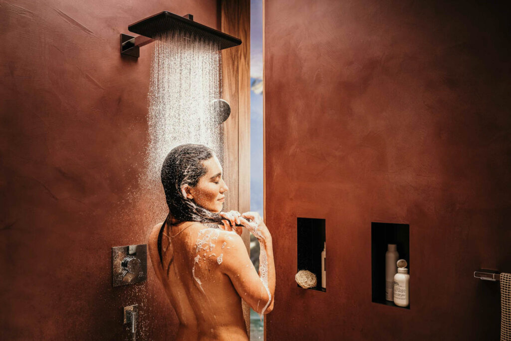Werbefotografie zeigt dunkelhaarige Frau genießt eine Dusche unter einer Hansgrohe-Dusche in einem roten Design-Badezimmer – Werbefoto mit Blick auf Rücken und Gesicht, stilvolle Bad- und Lifestyle-Inszenierung von Werbefotograf-Leipzig